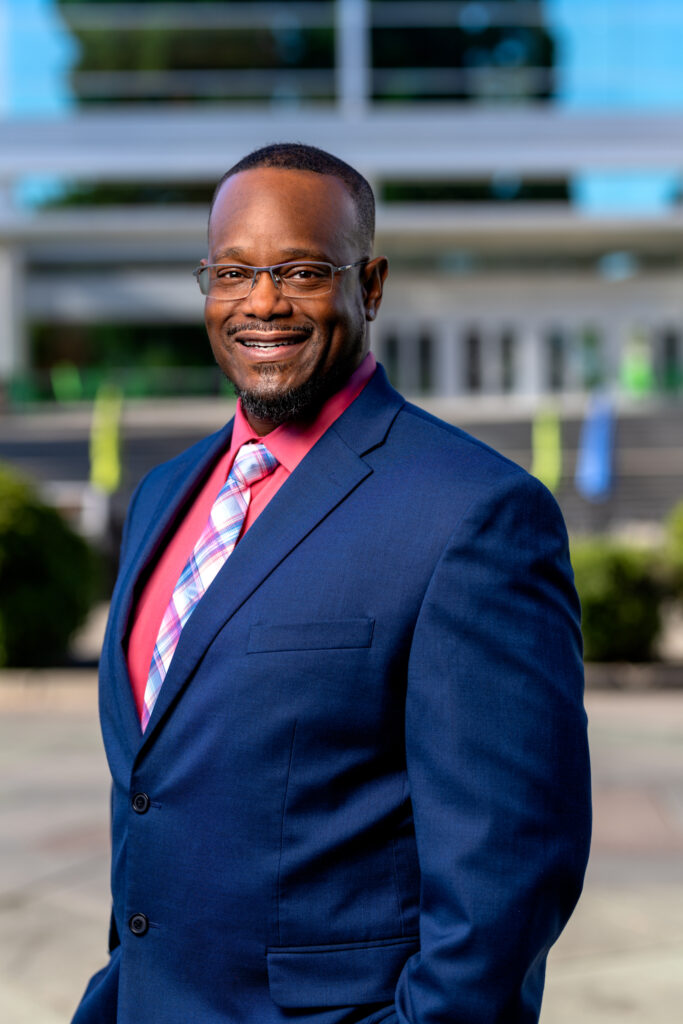 Titonian Wallace Sr. in a navy blue suit, pink shirt, and plaid tie stands outdoors, smiling confidently at the camera. The background is blurred, showing steps, greenery, and a modern building.
